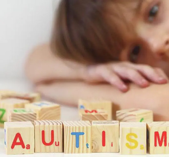 A young child rests their head on folded arms behind wooden alphabet blocks spelling “autism”